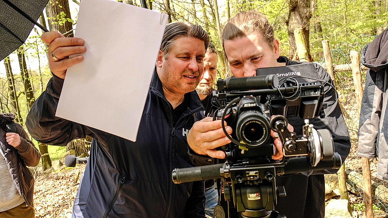 Ein Kameramann steht im Wald hinter einer Kamera und stellt diese ein. Ein Assistent hält ein weißes Blatt zum Weißabgleich in der Hand.